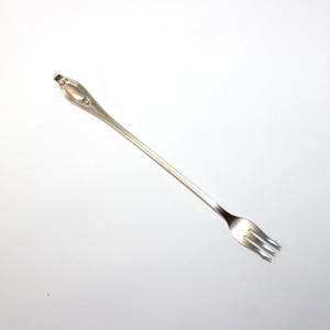 May include: A long-handled silver-toned cocktail fork featuring a slender stem and a decorative, oval-shaped embossed pattern on the handle. The fork has three thin, pointed tines and a polished metallic finish that reflects light across its surface.