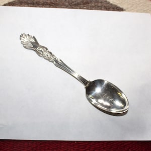 Peut inclure: Une cuillère en argent avec un manche orné. La cuillère est sur une feuille de papier blanche.
