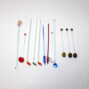 May include: A collection of decorative glass stir sticks with various colorful embellishments. The stir sticks have clear stems and are adorned with red, orange, green, blue, and gold accents. Some have gemstone-like toppers.