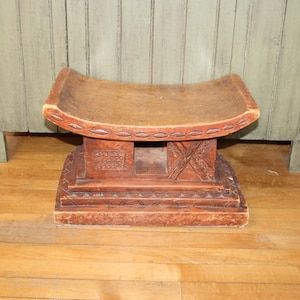 May include: A carved wooden stool with a curved seat and a detailed base. The stool is made of reddish-brown wood and features intricate carvings on the base and sides. The stool is approximately 12 inches long, 8 inches wide, and 10 inches tall.