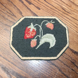 May include: An octagonal rug coaster featuring a dark green background with a central design of red strawberries, light green leaves, and thin stems. The piece is finished with a cream-colored border that outlines the eight-sided shape.