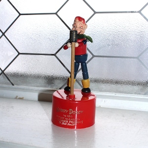 May include: A vintage Howdy Doody toy figure. The figure is red, blue, and yellow, holding a microphone. The toy is mounted on a red cylindrical base with the words "Howdy Doody" printed on it. The background is a window with a diamond pattern.