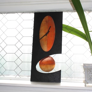 May include: A rectangular black wall clock featuring a vertical oval face with a gradient of orange and copper tones. The clock has thin black hands and a circular pendulum in a matching orange and copper finish suspended within a cutout section.