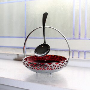 Peut inclure: Un sucrier en argent avec un insert en verre rouge et une cuillère en argent assortie. Le bol a un design décoratif ajouré autour du bord.