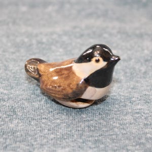 Puede incluir: Pequeña figura de pájaro de cerámica. El pájaro tiene cabeza negra, cuerpo marrón y detalles blancos. La figura está sobre una superficie azul claro.