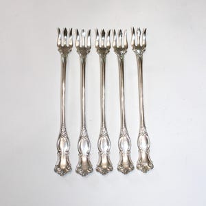 Puede incluir: Un juego de cinco tenedores de cóctel de metal plateado, cada uno con un mango largo y delgado con un diseño de volutas en relieve en la base y tres dientes afilados y puntiagudos en la parte superior para pinchar alimentos.