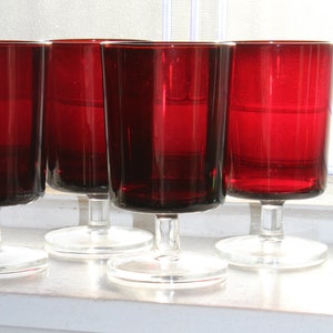 Pode incluir: Quatro taças de vidro vermelho com hastes e bases transparentes. As taças estão cheias de um líquido vermelho e estão dispostas em fila sobre uma superfície branca.