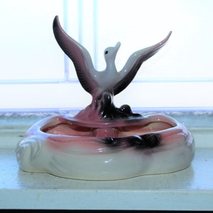 Puede incluir: Un plato de cerámica para baratijas con un esmalte rosa y negro. Un pájaro blanco con detalles negros está posado en la parte superior del plato.