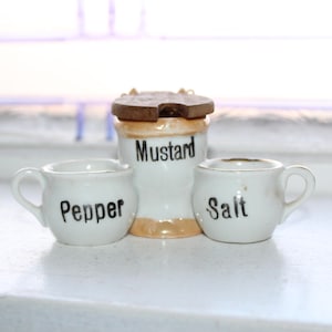 Puede incluir: Salero, pimentero y mostaza en cerámica blanca. El tarro de mostaza tiene una tapa de madera. Los tarros están etiquetados como "Sal", "Pimienta" y "Mostaza".