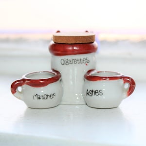 Puede incluir: Un juego de tres recipientes de cerámica con tapas rojas. Los recipientes están etiquetados como "Cigarrillos", "Cerillas" y "Ceniza".