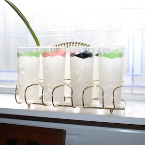 Pode incluir: Conjunto de cinco copos de vidro fosco com faixas coloridas ao redor da parte superior, que estão em um suporte de metal dourado. Os copos são transparentes e têm um design de floco de neve.