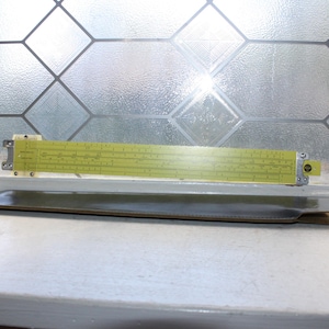 May include: A yellow slide rule with black printed scales and markings, featuring metal end brackets and a central sliding cursor. The instrument is positioned horizontally above a dark brown, flat, rectangular protective case.