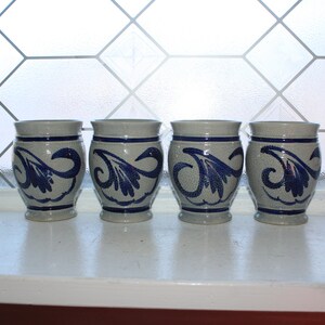 May include: Four gray ceramic mugs with blue and white floral designs. The mugs are lined up in a row on a white surface.