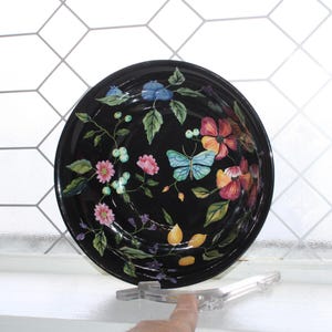 May include: A black ceramic serving bowl with a floral and butterfly design. The bowl features colorful flowers in shades of pink, orange, and blue, along with a light blue butterfly. Green leaves and stems complete the design.