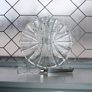 May include: A clear glass serving dish with a scalloped edge and a raised center design. A silver butter knife is placed in front of the dish.