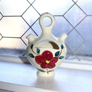 May include: Cream ceramic cruet with a red floral design. The cruet has a handle with a hole, two spouts, and a central opening. Green leaves and stems accent the flower. The cruet sits on a white surface.