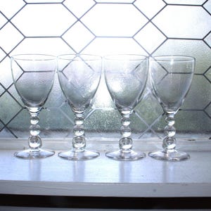 May include: A set of four clear glass goblets, each featuring a smooth, rounded bowl and a stem composed of three stacked glass spheres leading down to a circular base. The glasses are arranged in a row, showcasing their transparent material.