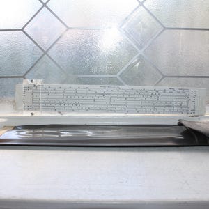 May include: A white plastic slide rule with black numerical markings and a clear sliding cursor, positioned horizontally above a long, flat, dark brown protective case made of a smooth, flexible material with a folded flap closure.