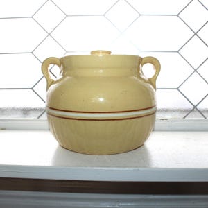 May include: A vintage, yellow-toned ceramic jar with a lid and two handles. The jar features a brown and white stripe around the middle. The jar is sitting on a white surface in front of a window.