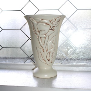 May include: Cream-colored ceramic vase with a flared top and a pedestal base. The vase features a raised floral design with brown accents. The background includes a window with a geometric pattern.
