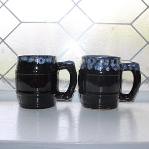 May include: Two black ceramic mugs with a barrel design and large handles. The rims have a blue and white speckled glaze. The mugs are displayed on a white surface in front of a window with a geometric pattern.