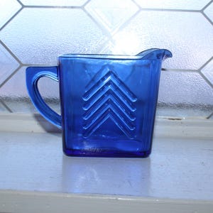 Puede incluir: Jarra de vidrio azul cobalto con forma cuadrada y asa. La jarra presenta un diseño decorativo de chevrón en la parte delantera. Está colocada sobre una superficie blanca frente a una ventana.