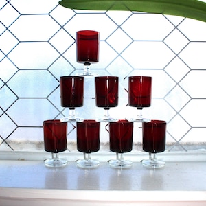 May include: A pyramid of eight ruby red wine glasses with clear stems and bases. The glasses are arranged on a white windowsill in front of a window with a geometric pattern. A green leaf is visible in the background.