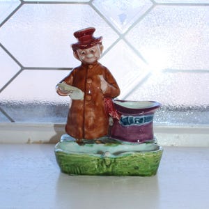May include: A ceramic figurine of a man in a brown coat and red hat, holding a paper. Beside him is a purple and blue top hat, all on a green base. The figurine is set against a window.