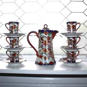 Puede incluir: Un juego de té decorativo con una tetera adornada, con asa y boquilla rojas, y varias tazas y platillos apilados. El juego tiene una base blanca con detalles rojos, azules y dorados. El fondo es una ventana.