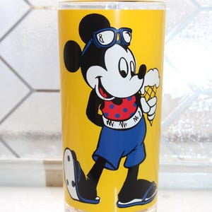May include: A clear glass tumbler featuring a bright yellow background with a graphic of Mickey Mouse wearing blue sunglasses, a red shirt with blue polka dots, and blue shorts, while holding a vanilla ice cream cone in a waffle cone.