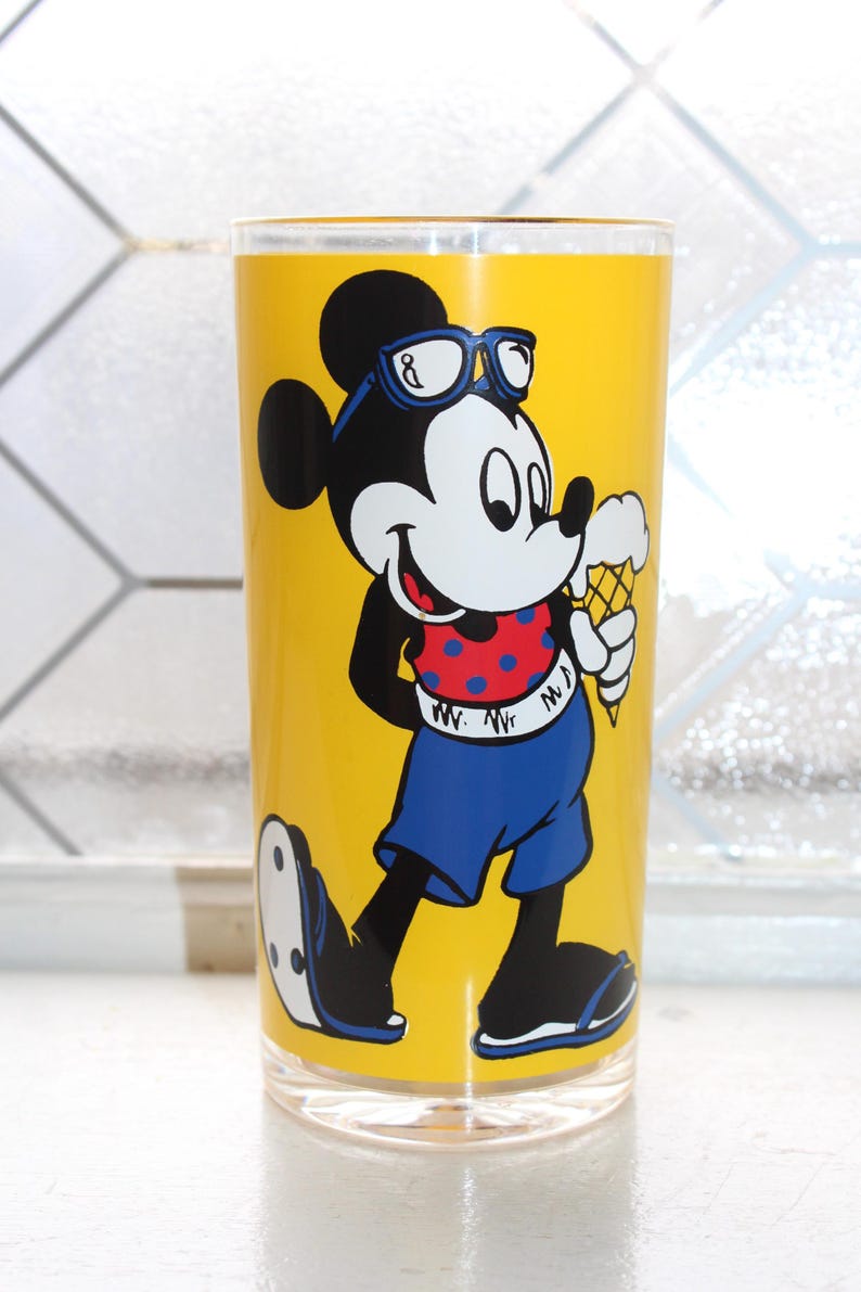 May include: A clear glass tumbler featuring a bright yellow background with a graphic of Mickey Mouse wearing blue sunglasses, a red shirt with blue polka dots, and blue shorts, while holding a vanilla ice cream cone in a waffle cone.