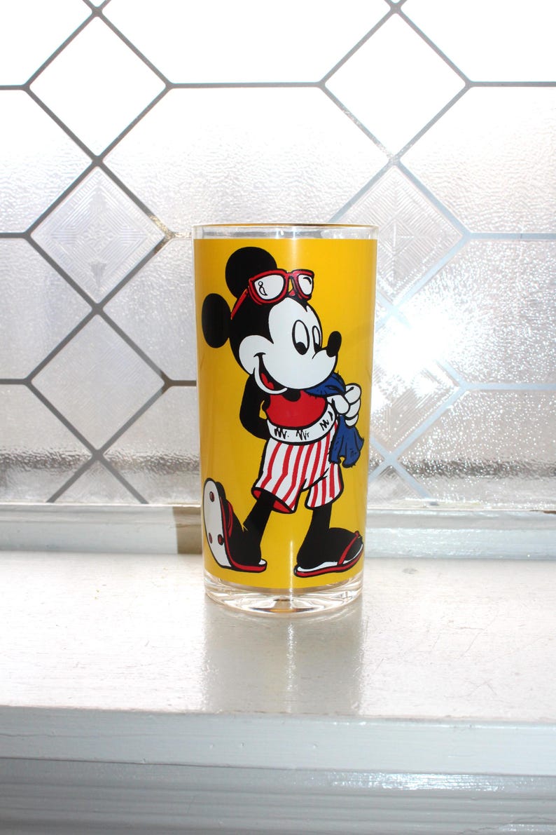 May include: A clear glass tumbler featuring a vibrant yellow background with a graphic of Mickey Mouse wearing red-rimmed sunglasses, a red top, and red-and-white striped shorts while holding a blue towel. The character is wearing black sandals.