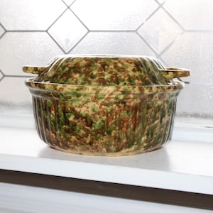 Può includere: Una pirofila in ceramica coperta con un motivo maculato verde, marrone e giallo. Il piatto ha due piccole maniglie ed è appoggiato su una superficie bianca. Lo sfondo è una finestra con un motivo geometrico.