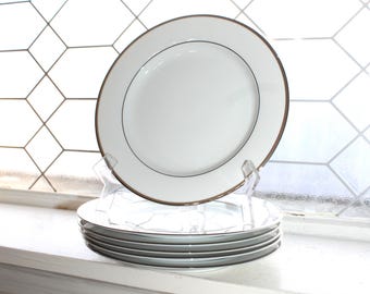 May include: A stack of white ceramic plates with a gold rim and a thin black line. One plate is displayed on a clear plastic stand. The plates are on a white windowsill with a geometric patterned window in the background.