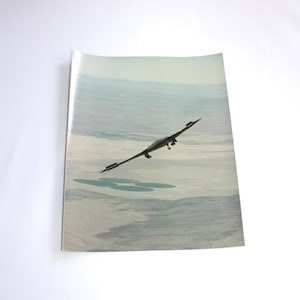 May include: A black aircraft with a long, narrow wingspan flies over a desert landscape. The aircraft has a distinctive, angular design and is flying at a low altitude.