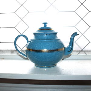 May include: A blue teapot with a gold band around the middle and on the lid. The teapot has a curved handle and spout. The background features a window with a geometric pattern.
