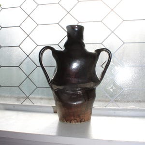 May include: A dark brown ceramic vase with two curved handles and a flared base. The vase has a glossy finish and sits on a white windowsill. The background features a window with a geometric pattern.