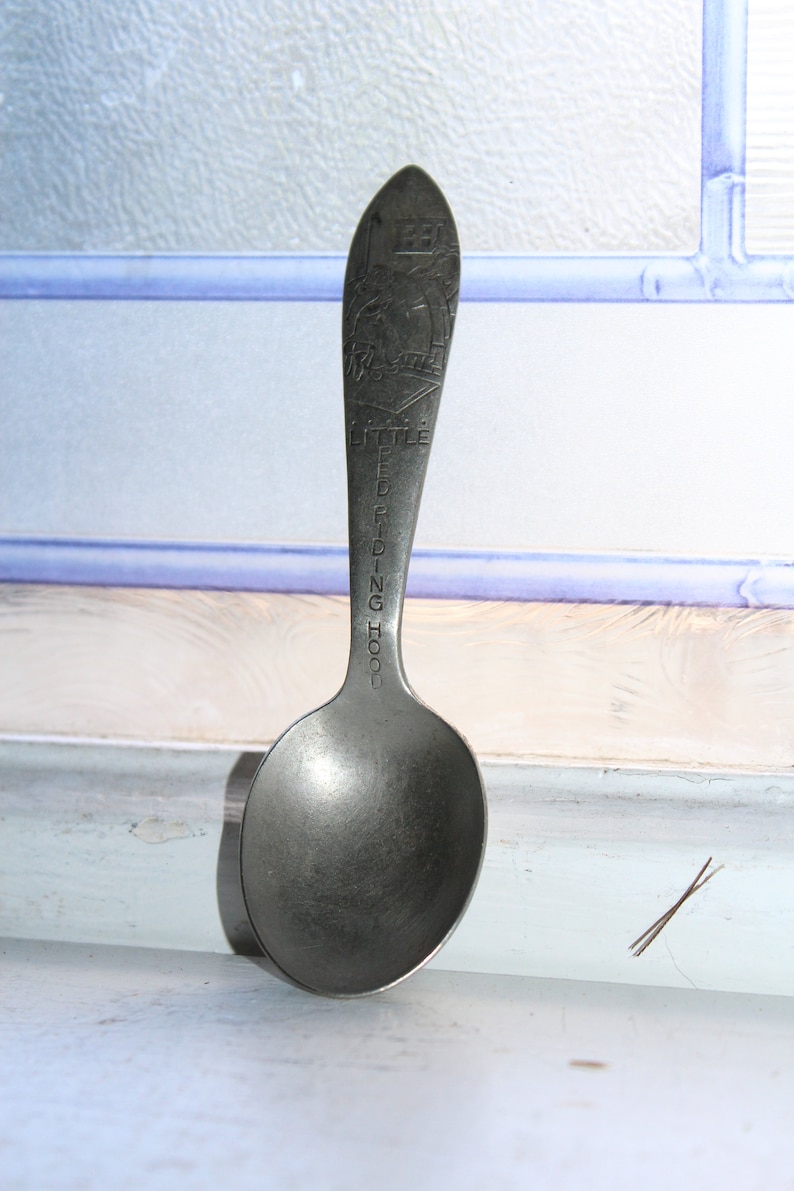 May include: A silver spoon with a rounded bowl and a long, slender handle. The handle is engraved with the words "Little Red Riding Hood".