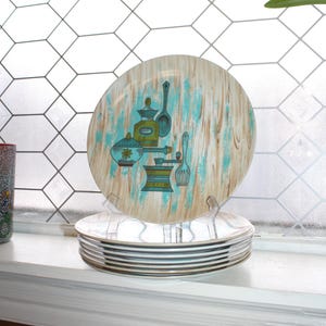 Op de afbeelding: Een stapel borden in vintage-stijl met een retro keukengerei-ontwerp. De borden hebben een lichte houtnerf achtergrond met turquoise en bruine accenten. De borden worden tentoongesteld op een witte vensterbank.