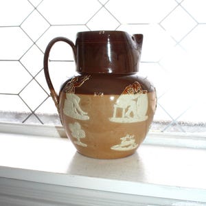 May include: A brown and tan ceramic pitcher with a handle and spout. The pitcher features white relief designs of figures, trees, and deer. The top and handle are a darker brown, while the body is tan. It sits on a white windowsill.