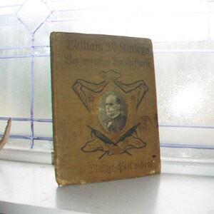 May include: A vintage book with a tan cover featuring a portrait of a man in a frame. The book's title is in gold lettering, with additional text and illustrations. The book is displayed on a white windowsill.