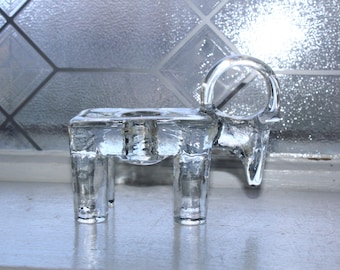 May include: Clear glass candle holder in the shape of a ram. The candle holder has a rectangular base with four legs and a circular horn detail. The glass is transparent, allowing light to pass through. The background is a window with a diamond pattern.