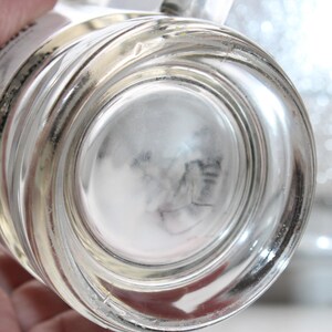 May include: A clear glass mug with a thick, circular base and a smooth, transparent body. The bottom of the mug features a recessed center, and the side wall shows a partial white label with black text visible through the glass material.