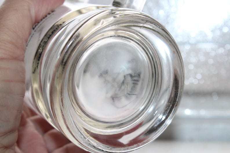 May include: A clear glass mug with a thick, circular base and a smooth, transparent body. The bottom of the mug features a recessed center, and the side wall shows a partial white label with black text visible through the glass material.
