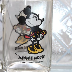 May include: A clear glass mug featuring a graphic of Minnie Mouse wearing a red and yellow polka dot dress and yellow shoes. The text MINNIE MOUSE is printed in black below the character, followed by the text &copy;Walt Disney Productions.