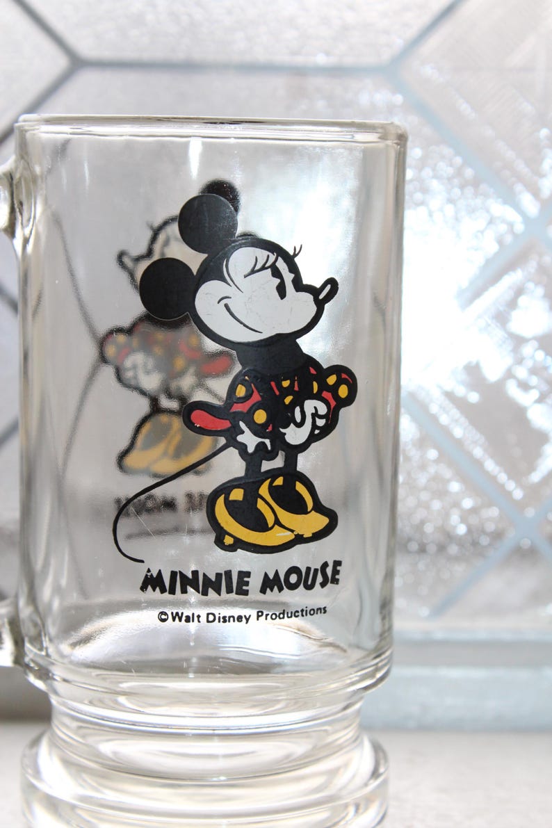 May include: A clear glass mug featuring a graphic of Minnie Mouse wearing a red and yellow polka dot dress and yellow shoes. The text MINNIE MOUSE is printed in black below the character, followed by the text &copy;Walt Disney Productions.