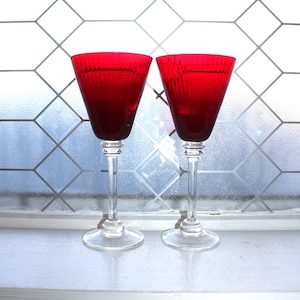 Könnte beinhalten: Zwei rubinrote Weingläser mit klaren Stielen und Böden. Die Gläser haben ein strukturiertes, geripptes Design auf der Schale. Sie stehen vor einem Fenster mit einem geometrischen Muster.