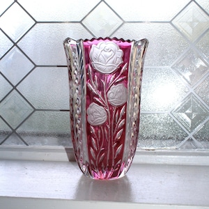 May include: A clear glass vase with a vibrant pink interior. The vase features a floral design with white roses and leaves. The vase is sitting on a white windowsill, with a frosted glass window in the background.