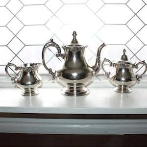 May include: A silver tea set, including a teapot, creamer, and sugar bowl, is displayed on a white windowsill. The set has ornate handles and decorative detailing. The background features a window with a geometric pattern.