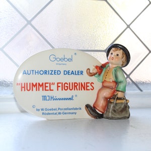 May include: White oval sign with blue and red text that reads "Goebel Germany Authorized Dealer "Hummel" Figurines M.I. Hummel by W. Goebel, Porzellanfabrik Rödental, W-Germany". A figurine of a boy with a black hat and a brown suitcase is standing on the sign.